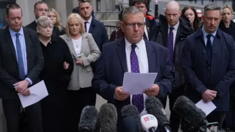 PA Media Gary Furlong, the father of victim James Furlong, watched by his wife Jan, and other victims' family members talks to the press outside the Old Bailey, central London, after the inquest concluded in April