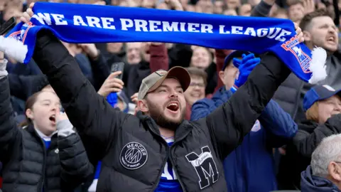 PA Media Ipswich Town fans can be seen cheering at a match, many of them waving their hands in the air. A young man is in the centre of the frame, wearing a black puffer jacket over a blue replica Ipswich shirt, a stone-coloured baseball cap and is holding a blue flag reading: "We are Premier League".