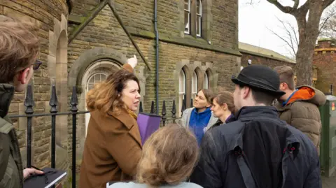 A woman in a brown coat points towards an old brick building. She is speaking to a crowd of people.