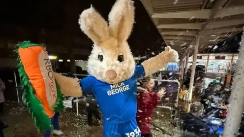 Richard Stangward Peterborough United mascot, a white rabbit wearing the Posh blue jersey, holding an orange carrot cheering at the lights switch-on event.