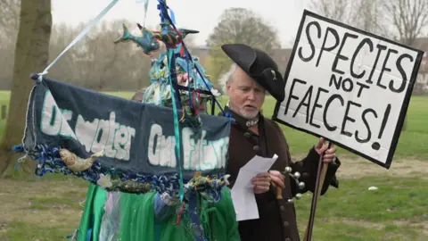 A man dressed in a brown coat and an old black hat, sat on his head at a jaunty angle, is holding a small tambourine and a placards which says 'Species not Faeces'. He is stood next to another person in a green and blue costume who is holding a banner that says 'Our water. Our future'