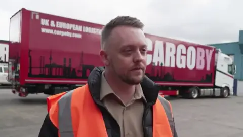 Tom Ingall/BBC A man with short blonde hair wearing a black jacket a beige shirt stands in a yard in front of a red truck