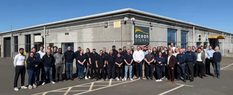 Ocean Signal Several men and women smiling smiling and posing for the photograph in a car park outside a single storey office building.
