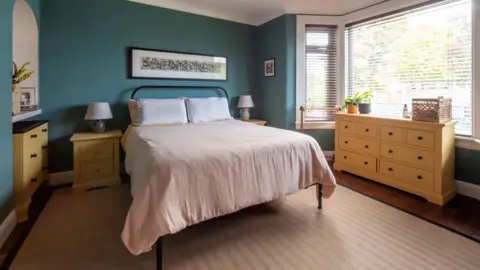IWC Media A calm, neatly arranged bedroom features deep teal walls, a central bed with neutral bedding, and matching light wood bedside tables and drawers.
Large bay windows with blinds let in natural light, complementing the warm tones of the wooden furniture and soft-textured rug.