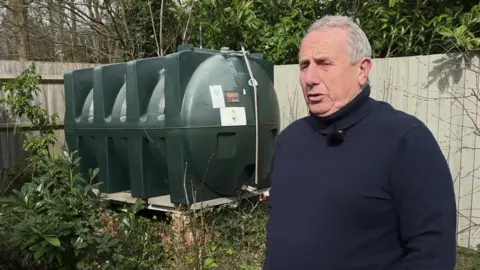 A man in a black turtleneck jumper stands in his garden. Behind him is a big green plastic tank filled with heating oil. 