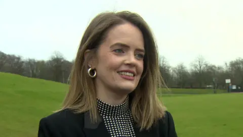 Trása Canavan. She has brown shoulder-length hair and blue eyes, wearing a black and white top and black blazer.