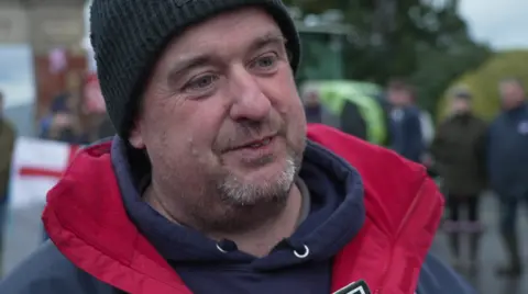 Christopher Marchment looking at the camera is wearing a navy hoodie, red gilet and a black beanie hat.