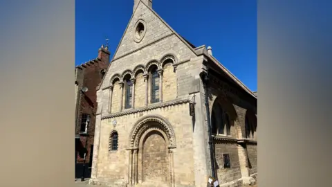 The Cromwell Museum The exterior of the Cromwell Museum in Huntingdon under a bright blue sky. It shows a medieval stone-built building with a bricked up door on the ground floor under a Norman-style arch, a row of largely bricked up arched windows above and rises to a point.