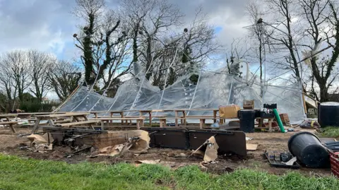 BBC A damaged outdoor structure with a collapsed transparent canopy in a muddy area surrounded by bare trees. The frame of the canopy is bent and partially torn, with clear plastic sheets hanging loosely. Several wooden picnic benches are arranged underneath the damaged canopy. In the foreground, broken furniture, scattered debris, and large black plastic containers are visible on the ground.