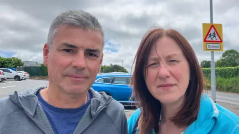 Michael and Veronica McLaughlin are standing side-by-side. Michael is wearing a grey hoodie and blue shirt. Veronica is wearing a light blue hoodie. They are both standing in a car park.
