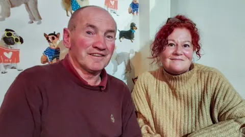 John Kelly with shaved hair wearing a brown polo top and jumper and Carole Kelly with auburn curly hair and black glasses resting on her head wearing a beige roll neck jumper. They are sitting in a pub and both are smiling.