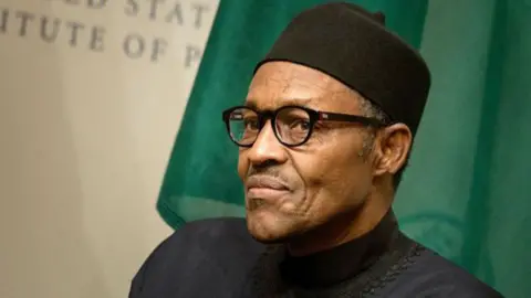 Muhammadu Buhari sits in front of a Nigerian flag and signs that say United States Institute of Peace. He wears a black top, black cap and glasses.