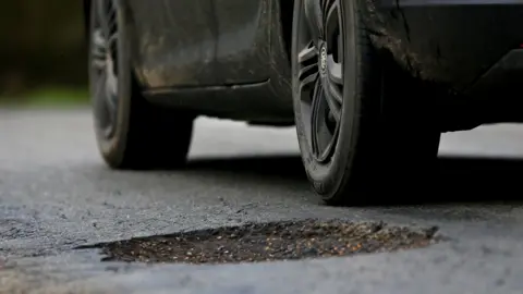 PA Media Close-up photograph of a muddy black car on a road near a large pothole.