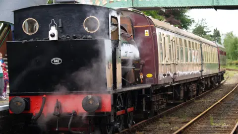 A black steam engine at a station connecting up to three burgundy-coloured carriages. 