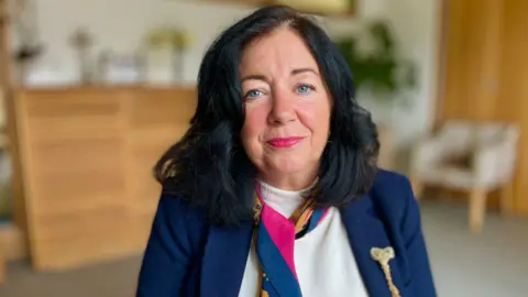Anne-Marie Hoare looking into camera in a room at the Arthur Rank Hospice. She has long, dark hair and is wearing a navy suit, white jumper and pink and navy scarf.
