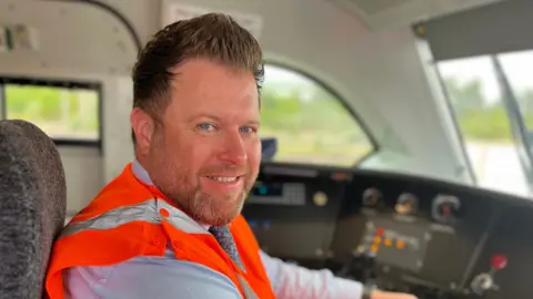 Emma Baugh/BBC Mr Garner is inside the new locomotive and appears to be driving it. He is looking at the camera and smiling. He is wearing an orange high vis vest with a suit and tie. He has blue eyes, some facial hair and short brown hair that has been styled to stick up slightly at the top of his head. In front of him is a control panel with dials and buttons used to control the locomotive. 