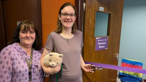 Anna Brader and Alisha smiling at the camera whilst Alisha holds scissors next to a purple tape with a door saying 'welcome to the sensory room'