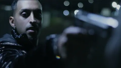 Kay S. Ubhi/Miracle Media A scene from a film in which a man in black clothing points a gun at someone unseen. His face is in shadow and lights are behind him.