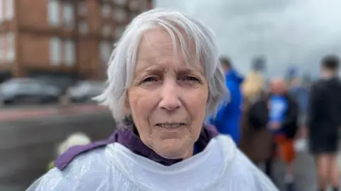 Maureen Douglass looking right at the camera. She has grey hair and is wearing a white t-shirt over a purple jacket.