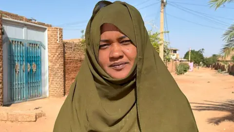 Ken Mungai / BBC Asma Mubarak Abdel Karim di Abaya Khaki Green Khaki Dark melihat kamera saat dia berdiri di jalan berdebu di Haji Yusuf, distrik Khartoum, Sudan - Maret 2025