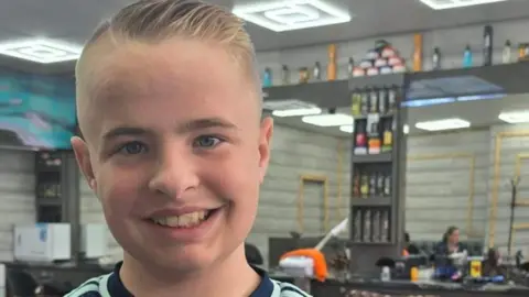 Cancer Research UK A smiling young boy with short blonde hair stands in a barbers shop.