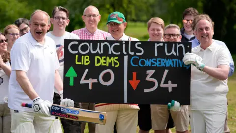 Sir Ed Davey pictured with a group of councillors who are wearing cricket gear. Sir Ed is holding a cricket bat up in the air, while holding a score board in the other which shows the Lib Dem's have gained 40 seats and the Conservatives have lost 34.