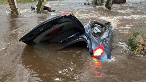 Hampshire and Isle of Wight Fire and Rescue Service A half-submerged grey hatchback car tilted on its side in floodwater. The lights are on and the boot is open wide.