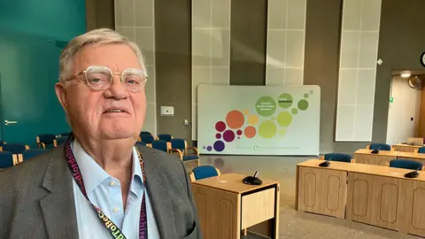 Amy Holmes/BBC A picture of councillor Steve Owen who is stood in the council chambers at Central Bedfordshire Council. He has a number of desks and chairs behind him and is wearing a light blue shirt, a dark grey jacket and has glasses on.