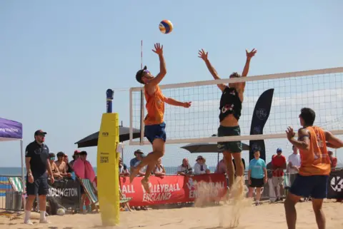 Jon Cornish/Volleyball England A beach volleyball match taking place on a sunny day. Two players - one in an orange vest and one in a dark vest - are both jumping at the net, reaching up toward a volleyball. Another player in an orange vest is standing nearby. Spectators are watching from behind a barrier, with beach umbrellas, banners and flags visible in the background. The sea and clear blue sky are also visible behind the court.