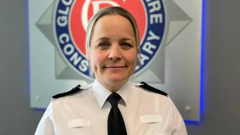 Deputy chief constable of Gloucestershire Police, Katy Barrow-Grint. She is wearing a white shirt with black epaulettes and a black tie. She has blonde hair tied up into a pony tail. She is standing in front of a Gloucestershire constabulary sign.
