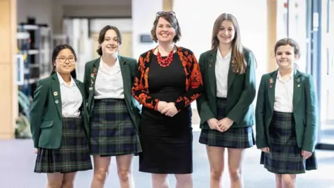 Principal Daniele Harford-Fox wearing a black dress and floral cardigan. She has short dark hair. Two female students are standing either side of her. 