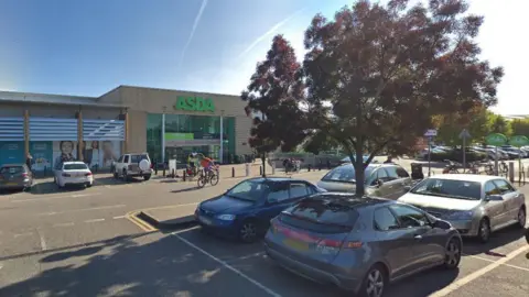 Google The outside of Asda, showing a large brick building with the Asda logo above the entranceway. In the foreground are parked cars and a tree.