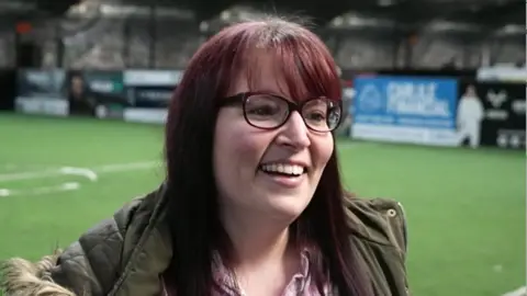 Hayward-Bannister stands on an indoor football pitch. She is wearing a khaki‑green coat with a fur‑trimmed hood and a light pink top underneath. Her hair is long, straight, and dark with a reddish tint. Behind her, the artificial turf stretches across the facility, and advertising boards line the walls.