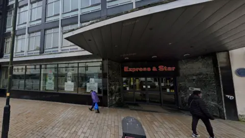 Google A google streetview picture showing the outside of the former Express and Star newspaper office. There is a person wearing a purple coat and carrying a blue plastic bag walking past the window and a person in a black padded coat standing on the opposite side of the pavement 