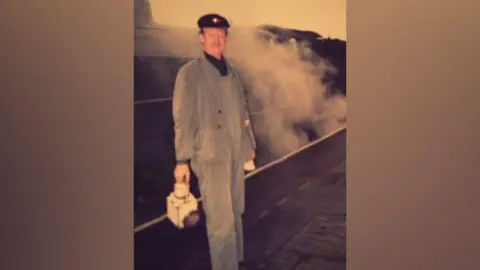 Supplied Richard Law is dressed in light blue overalls and is holding a light. He is stood on a platform next to a stream train