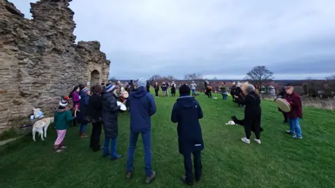 Charlotte Knaggs More than 20 people stand in a circle near a ruined castle drumming. They are joined by one dog