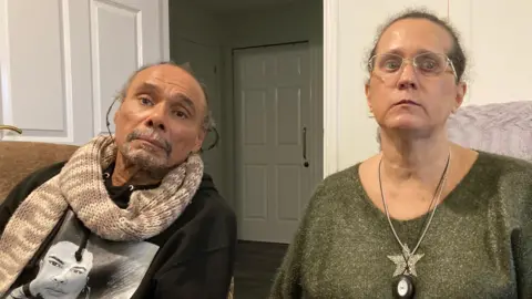 A man and a woman sit side by side indoors. The man wears a dark hoodie with a printed portrait and a knitted scarf. The woman wears glasses, a green jumper, and a necklace.