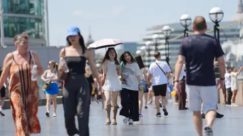 People walk through London in a heatwave, one carrying a bottle of water and another using an umbrella as a sunshade.