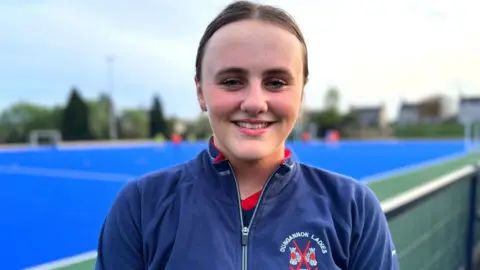 A girl smiles into the camera. She has brown hair, which is tied back. She is wearing a navy Dungannon Ladies hockey top. She is standing in front of a blue hockey pitch.
