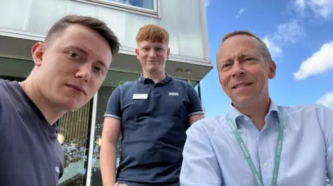 Three men, all in blue tops, pose in front of a camera for a selfie. 