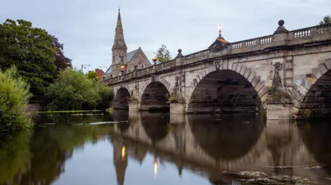 The River Severn running over a large stone bridge with several arches. There is a church in the background with a spire, and green trees and bushes on the river side