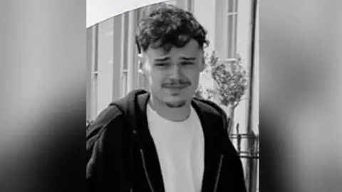 Police handout Black and white photograph of William Sullivan. He has brown curly hair and stubble. He is wearing a white t-shirt and black hoodie outside a building.