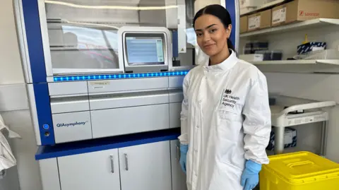 A woman with brown hair in a ponytail smiles at the camera wearing a white lab coat and blue rubber gloves. She stands in front of a machine that processes biological samples in a laboratory