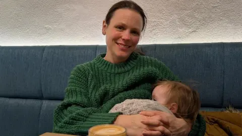 Alexis Fullard A woman in a chunky knit green jumper loops her arms round an infant who is snuggled into her chest. The woman has dark hair tied back and smiles as she looks at the camera. She is sitting on a blue fabric bench and has a flat white in front of her on a table.
