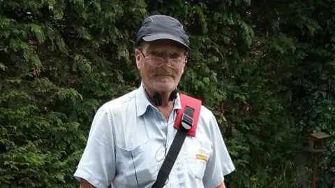 Keith is standing in a garden.
There are fir trees and a table in the background.
He is wearing a light blue, short sleeved Royal Mail shirt, and carrying a letter bag over his left shoulder.
He is wearing glasses.