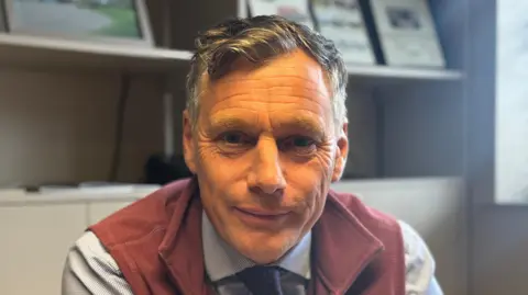 George King/BBC A head and shoulders image of Oliver Johnson. He is wearing a red sleeveless top over a shirt with a tie. He is looking directly into the camera. 