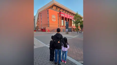 Three young children have their backs turned to the camera as they stand arm in arm looking on at the Anfield stadium.