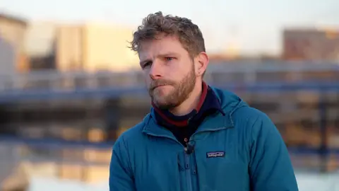Image of Josh Doble from Community Land Scotland. He has short, light brown hair and beard and moustache. He is wearing a red and navy scarf under a zipped blue waterproof jacket  