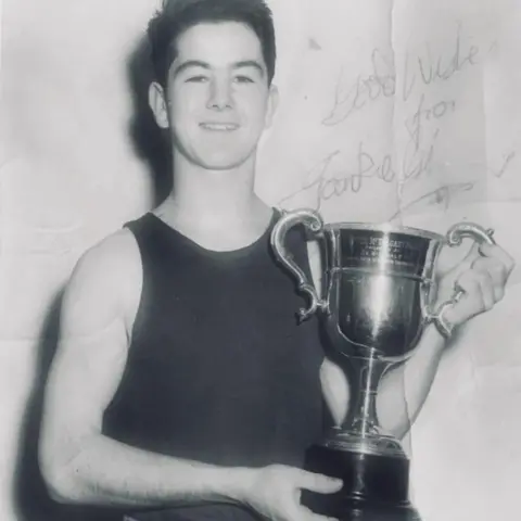 Paul Gilfeather Una fotografía antigua en blanco y negro de un joven Frank Gilfeather con una camiseta oscura y sosteniendo un trofeo.