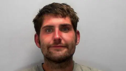 A police mugshot of Jack David Anthony Merry. He has short brown hair and a brown beard and wears a grey jumper.
